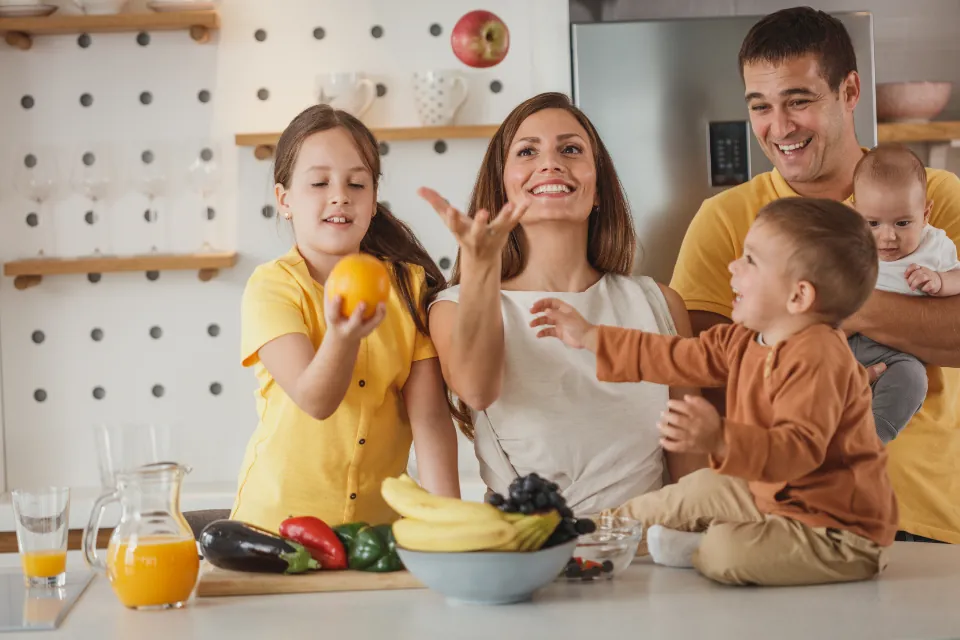 A family laughing together in a bright kitchen, the daughter holding an orange